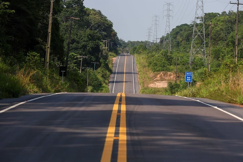 “Desde janeiro faço essa rota e percebi a diferença. Antes era só buraco, hoje está mil vezes melhor. Consegui adiantar quase uma hora de viagem.” O relato é de Janes Mário, 25 anos, representante comercial que percorre com frequência o trecho entre Rio Preto da Eva e Itacoatiara pela rodovia AM-010. Esse é um dos vários benefícios dos investimentos do Governo do Amazonas, na vida da população, com o Programa Asfalta Amazonas, que engloba ações de pavimentação e revitalização em ruas, ramais e rodovias na capital e no interior. A rotina de Janes Mário na rodovia AM-010 é semelhante à de centenas de pessoas que, no último mês, passaram a experimentar uma rodovia mais rápida, segura e confortável. A mudança na vida de quem depende diariamente da estrada é resultado da maior intervenção já realizada na AM-010 em quatro décadas. A obra, financiada integralmente pelo Governo do Amazonas, marca um novo ciclo de investimentos em mobilidade e infraestrutura no estado, que vem sendo reforçado com ações simultâneas em diferentes regiões, tanto em vias estratégicas quanto em áreas urbanas e rurais. “Dar melhores condições de infraestrutura é dar mais dignidade para as pessoas. A AM-010 é a estrada mais importante do Amazonas e desde que assumimos o governo, firmamos o compromisso de melhorar as condições da rodovia e hoje vivemos um novo momento”, disse Wilson Lima. Inaugurada no dia 15 de novembro, deste ano, pelo governador Wilson Lima, a primeira etapa da modernização da AM-010 entrega 142 quilômetros completamente reconstruídos entre o KM 110 e o KM 252, no trecho que liga Rio Preto da Eva a Itacoatiara. Com investimento de R$ 515,7 milhões, a rodovia possui 250,40 quilômetros no total e é um dos principais eixos estruturantes do Amazonas, essencial para o transporte de insumos e mercadorias, o escoamento da produção agrícola, o acesso a serviços públicos e a circulação de trabalhadores, estudantes e turistas. O ritmo acelerado permitiu que a estrada fosse modernizada em aproximadamente quatro anos, em contraste com obras semelhantes, como a AM-070, iniciada em 2013 e concluída apenas em 2021, na atual gestão estadual, responsável por finalizar quase 60% dos serviços em três anos. A modernização da mais importante rodovia estadual representa avanço significativo sobre mais de 2 milhões de amazonenses com redução do tempo de viagem, que antes chegava a seis horas, para cerca de três; maior segurança, diminuição de custos de manutenção de veículos e melhoria direta no deslocamento diário de quem depende da via. A obra traz benefícios diretos para Manaus, Rio Preto da Eva e Itacoatiara, e reflexos positivos em municípios como Itapiranga, São Sebastião do Uatumã, Silves, Urucará e Urucurituba. A modernização também impulsionou o mercado local, gerando mais de 10 mil empregos diretos e indiretos ao longo de sua execução. Mais obras Paralelamente à primeira etapa da AM-010, o Governo do Amazonas avança com o Programa Asfalta Amazonas, que reúne ações de pavimentação e revitalização em ruas, ramais e rodovias tanto na capital quanto no interior. A iniciativa, realizada com execução direta do Estado ou por meio de convênios com prefeituras, já alcança 38 municípios, levando recapeamento, construção de calçadas, meio-fio, sarjetas e nova sinalização viária. No município de Coari (a 363 quilômetros de Manaus), o programa segue com frentes de obras em diferentes localidades, totalizando 16,67 quilômetros de novas vias e R$ 25 milhões em investimentos. Os serviços em andamento contemplam bairros como Chagas Aguiar, Liberdade e União, além de trechos previstos em convênios já firmados entre o Governo do Estado e a prefeitura. Moradora do bairro Chagas Aguiar, a pensionista Valdelira Gama, 62, destacou a mudança na mobilidade e na qualidade de vida após a chegada das obras de pavimentação. “Antes quase ninguém podia andar com tanto buraco nessa rua. Então, para mim, isso é um grande milagre e um grande privilégio ver as obras”, agradeceu. Rodovias Desde 2019, seis rodovias estaduais já tiveram sua pavimentação concluída, totalizando 108,39 quilômetros e somando mais de R$ 791 milhões em investimentos. Outras quatro seguem em andamento, com 436,08 quilômetros e mais de R$ 1,34 bilhão, aplicados para garantir integração e trafegabilidade entre diferentes regiões. Ramais e estradas No interior, ramais e estradas também passaram por transformações significativas. Ao todo, 43 trechos já foram finalizados desde 2019, resultando em 248,85 quilômetros pavimentados e R$ 319,7 milhões em investimentos. Mais 21 obras seguem em execução, ampliando a malha com outros 265,96 quilômetros e recursos superiores a R$ 553 milhões, destinados a garantir trafegabilidade a agricultores, estudantes e famílias que dependem dessas vias para acessar serviços básicos. Sistema viário Nas áreas urbanas, 61 sistemas viários foram concluídos, somando 641,11 quilômetros de vias recuperadas com investimento superior a R$ 560 milhões. Outras 19 obras seguem em andamento, com 165,35 quilômetros e mais R$ 239,5 milhões aplicados em pavimentação, drenagem e urbanização. “Antes era só buraco. Hoje ganho quase uma hora de viagem”, usuário da AM-010 relata impacto da obra de modernização da rodovia